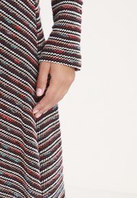 Main posée sur la hanche, portant une robe-pull tricotée rayée multicolore avec une texture côtelée, sur fond uni.