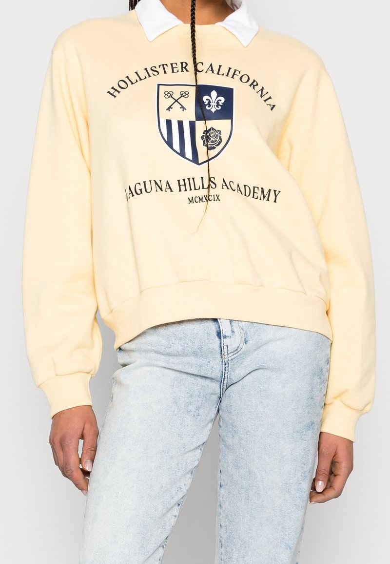 Person som bär en gul sweatshirt med texten "Hollister California Laguna Hills Academy" och ett vapen, tillsammans med ljusblå blekta jeans.