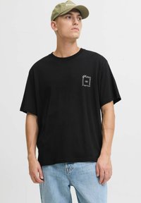 Jauns vīrietis valkā gaiši zaļu cepuri, melnu pārāk lielu T-kreklu ar baltu drukājumu un gaiši zilos džinsos, skatoties uz sāniem uz viendabīgas fona.