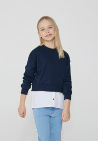 Marineblaues, kurzes Sweatshirt mit langen Ärmeln, gerippten Bündchen und rundem Halsausschnitt, über einem weißen Hemd mit Kragen und Knopfdetaillierung getragen.