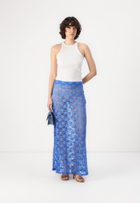 Jupe maxi en dentelle bleu clair superposée sur une doublure bleue unie, avec une texture lisse. Assortie à un débardeur blanc ajusté et à un petit sac bleu.