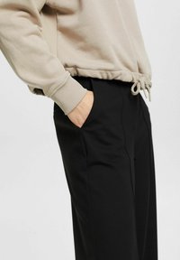 Beige sweatshirt med ribbade muddar och justerbar dragsko i nederkant, matchad med svarta byxor med sidofickor och en slät yta.