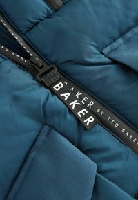 Doudoune bleue avec texture matelassée, dotée d'une fermeture zippée noire et d'un détail de label "BY TED BAKER" en blanc. Tissu lisse et brillant.