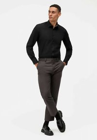 Camicia nera a maniche lunghe con bottoni, abbinata a pantaloni grigio scuro su misura. Scarpe nere robuste completano il look, su sfondo bianco.