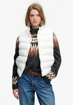 Ung kvinde med kort blondt hår har en hvid dynevest på over en mønstret mørk striktrøje og blå jeans med et flettet brunt bælte.