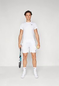 Hvit atletisk t-skjorte og shorts, svart Nike-logo på skjorten, holder en tennisracket og en gul tennisball, står på en nøytral bakgrunn.