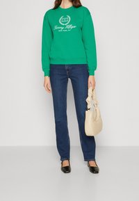 Felpa verde con logo bianco, abbinata a jeans blu scuro e scarpe nere slip-on. Borsa beige con un accento a fiocco. Outfit semplice e casual.