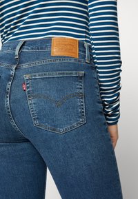 Tmavě modré džínové kalhoty s hnědou koženou značkovou náplastí, které mají zakřivený design zadní kapsy a červenou štítek na zadním švu.