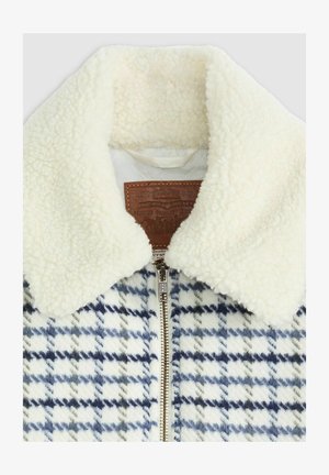 Veste doublée en polaire avec un motif pied-de-poule blanc et marine, dotée d'un col crème et d'un tag en cuir marron. Fermeture éclair sur le devant.
