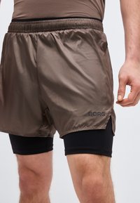 Άνδρας με καφέ φαρδιά σορτς πάνω από μαύρα εφαρμοστά συμπιεστικά σορτς, ορατός από τη μέση έως το μέσο του μηρού, στέκεται μπροστά σε απλό φόντο.