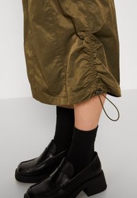 Jupe vert olive avec côté froncé ajustable et cordon, portée avec des chaussettes noires montantes et des mocassins compensés noirs.