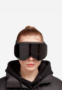 Žmogus su raudonais plaukais, vilkintis juodą džemperį su kapuce ir dėvintis per dideles, futuristines juodas akinius nuo saulės, priešais paprastą baltą foną.