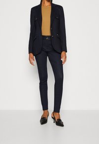 Námornicko modrý blejzr se dvěma předními kapsami a zlatými knoflíky, v kombinaci se světlým topem a tmavými skinny džínami, doplněný černými lodičkami na podpatku.