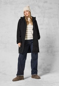 Manteau long noir avec capuche, bonnet beige côtelé, pull rayé crème et beige, jeans bleu foncé et chaussures brunes. Tenue décontractée d'extérieur.