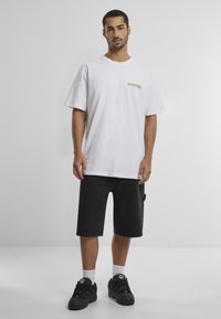 Camiseta de algodón blanca con un gráfico verde en el pecho, combinada con shorts de mezclilla negros y zapatillas negras. Calcetines blancos visibles.