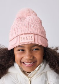 Lyserød strikket hue med en kunstig pelskvast, tekstureret kabelmønster og dekorative perler. Indeholder et mærket patch på manchet.