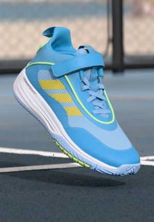 Blue athletic shoe with yellow stripes and green accents, suspended above a tennis court surface near white boundary lines.