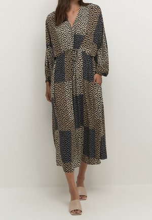 Femme portant une robe midi à manches longues et col en V avec un imprimé géométrique noir, beige et marron, et des mules à talons ouverts beige.