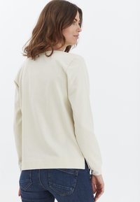 Camiseta de manga larga color beige claro con un ajuste relajado y aberturas en los laterales, hecha de una tela suave, combinada con jeans azul oscuro.