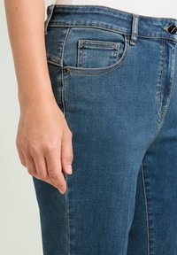 Donkerblauwe spijkerbroek met een gladde textuur. Voorzien van een voorzak, riemlussen en een knoopsluiting. Close-up van de zijkant.