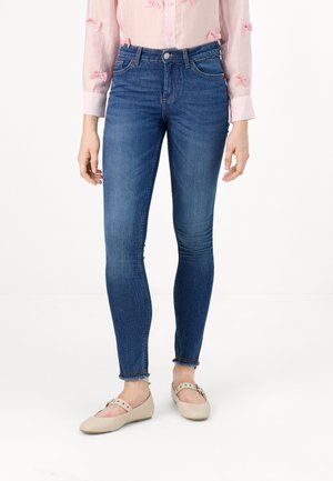 Jeans skinny de mezclilla en azul oscuro con una cintura alta, un desgastado sutil y con dobladillos sin terminar. Combinados con zapatos planos de color claro.