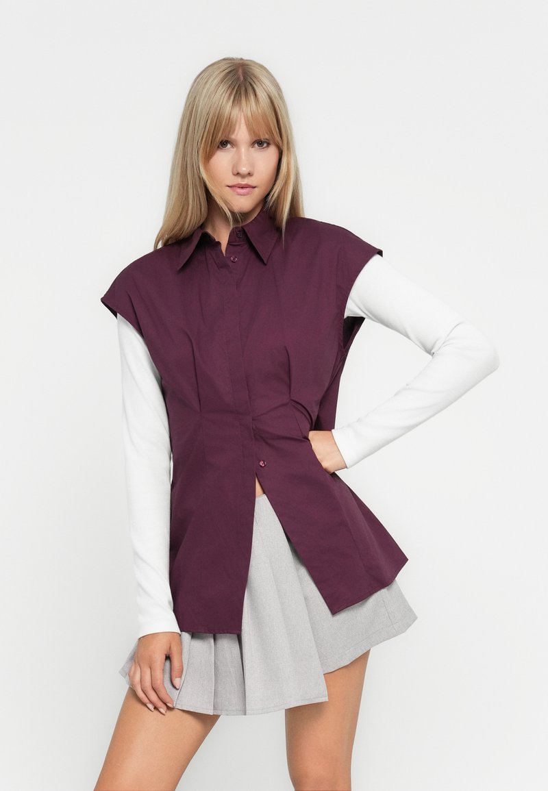 Bordeaux mouwloos overhemd met een kraag en knopen, gelaagd over een witte lange mouw top. Gecombineerd met een grijze geplooide rok.