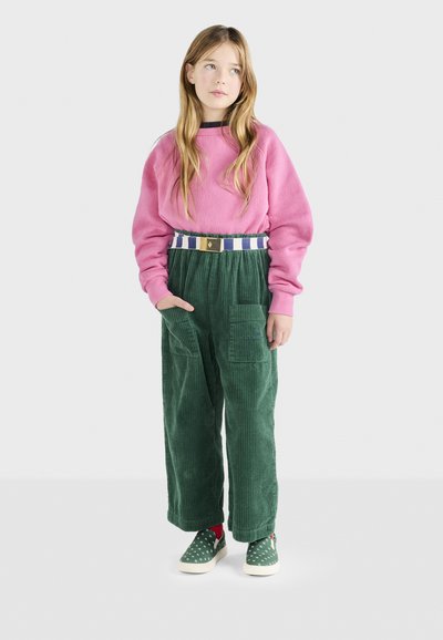 Rosa Fleece-Sweatshirt mit Raglanärmeln, grüne Cordhosen mit weitem Bein, gemusterter Gürtel und grüne Canvas-Slipper mit weißen Punkten.