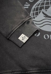 Musta collegepaita, jossa on resoroidut hihansuut ja kudottu merkkilappu logolla. Kankaalla on sileä pinta ja hienovarainen kiilto.