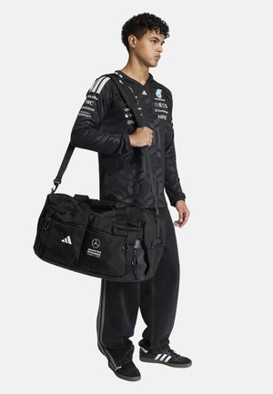 Joven vestido con chaqueta negra del equipo Mercedes AMG Petronas, pantalones negros, llevando un gran bolso negro de Adidas al hombro.