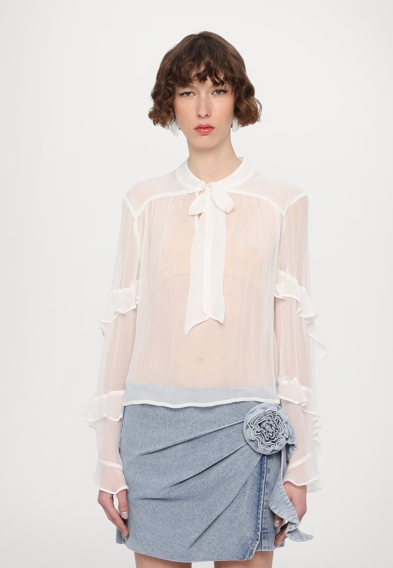 Gennemsigtig hvid bluse med volanger på ærmerne og en sløjfe ved kraven, parret med en blå denimnederdel med en blomsterdetalje.