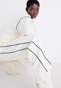 Survêtement crème avec des rayures noires sur les côtés, comprenant une veste zippée et un pantalon ample, associé à des baskets blanches épaisses.