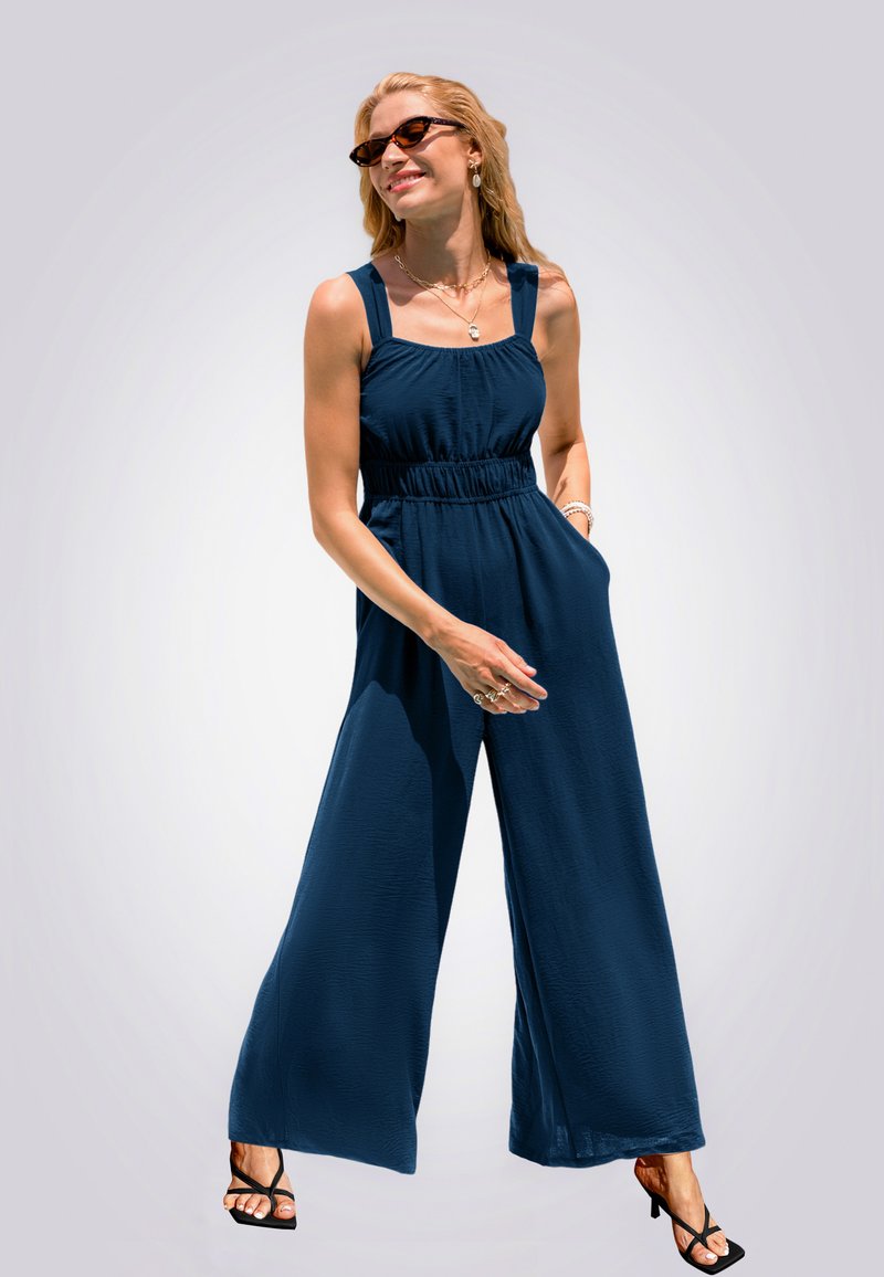Navyblauer Jumpsuit mit weitem Bein, elastischem Bund und breiten Trägern. Glattes Material mit Taschen, kombiniert mit schwarzen Riemchensandalen.