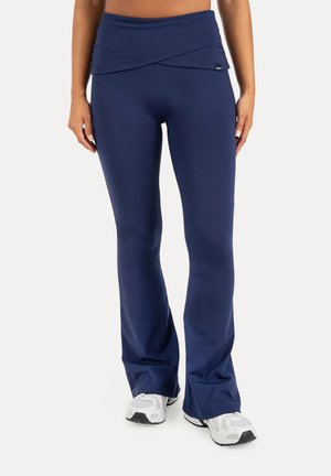 Mujer que lleva puestos leggings acampanados azul marino de talle alto y zapatillas deportivas blancas, de pie sobre un fondo blanco liso.