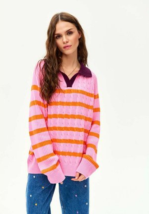 Młoda kobieta o długich, kręconych brązowych włosach, ubrana w różowo-pomarańczowy sweter w paski oraz niebieskie dżinsy z drobnym kwiatowym haftem.