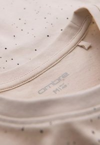 Lichtbeige t-shirt met zwart gespikkeld patroon. Ronde hals met een zachte textuur. Etiket leest "ombre M." Stiksel zichtbaar bij de kraag.