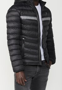 Black puffer jacket with a hood, featuring horizontal quilting, a silver accent stripe, and zippered side pockets.