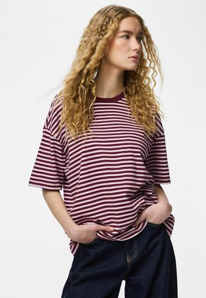 Jeune personne aux longs cheveux bouclés blonds portant un t-shirt rayé bordeaux et blanc et un jean foncé, mains dans les poches, regardant vers la droite.