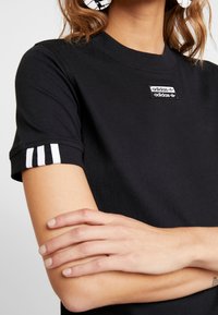 Svart bomull t-shirt med korta ärmar som har vita tredelade detaljer på manchetterna och en liten svart Adidas-logotyp på bröstet.