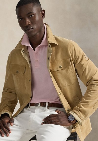 Νέος άνδρας που φοράει μπεζ μπουφάν πάνω από ροζ πόλο πουκάμισο και λευκό παντελόνι, καθισμένος με το ένα χέρι στη μέση και φοράει ρολόι χειρός.