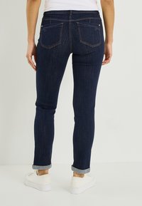 Jean en denim bleu foncé à coupe slim, ourlets retroussés et deux poches arrière avec des surpiqûres contrastées. Texture lisse et design décontracté.