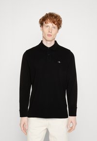 Black long-sleeve polo shirt in a smooth fabric with a buttoned collar and subtle logo detail on the chest, paired with light-colored trousers.