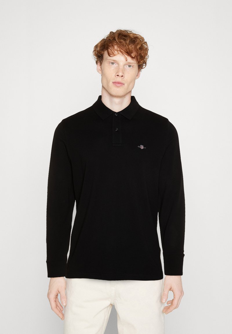 Black long-sleeve polo shirt in a smooth fabric with a buttoned collar and subtle logo detail on the chest, paired with light-colored trousers.
