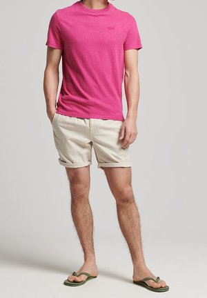 Mand iført en lyserød kortærmet t-shirt, beige shorts med opkrogede ben og grønne klip-klappere, stående med hænderne i lommerne.