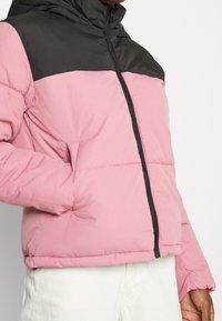 Giacca a vento gonfiabile con un corpo rosa e una parte superiore nera, dotata di chiusura a zip e tasche laterali. Il materiale sembra liscio e imbottito.