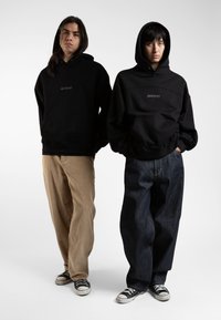 Black hoodies with logo, paired with beige corduroy and dark denim pants. Both models wear black Converse sneakers.