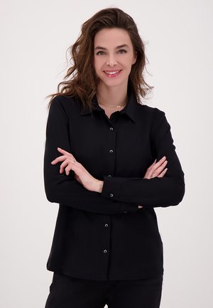 Zwarte overhemd met kraag, lange mouwen en een gladde textuur. Heeft bijpassende knopen en een relaxte pasvorm. Zelfverzekerd gedragen met gekruiste armen.