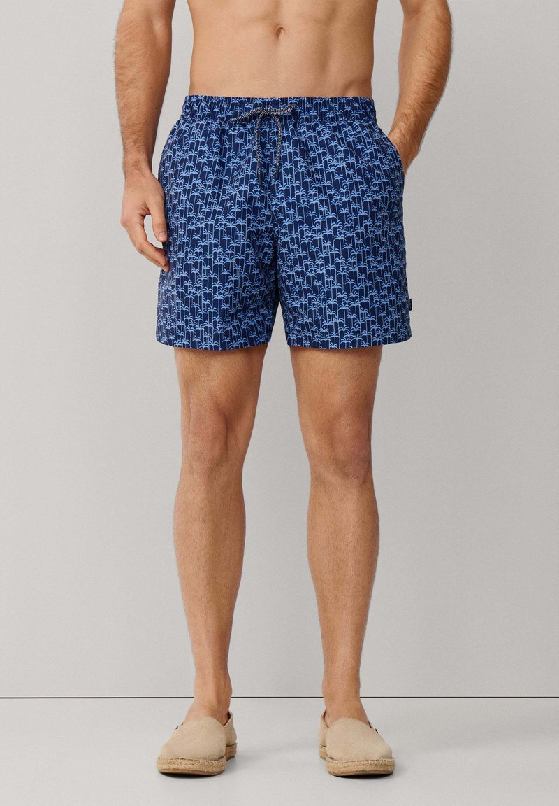Homme portant un short de bain bleu à motifs avec poches latérales et des chaussures beige à enfiler, debout devant un fond uni.