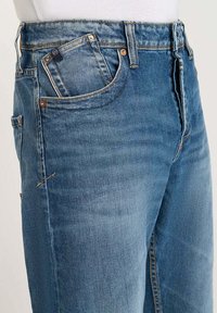 Jeans en denim bleu avec un design à jambe droite. Les caractéristiques incluent une poche avant, des coutures visibles et une légère décoloration sur le tissu.