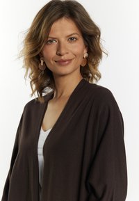 Bruine open voorkant cardigan over een witte top, met een relaxte pasvorm en zachte textuur, gecombineerd met beige ronde oorbellen.