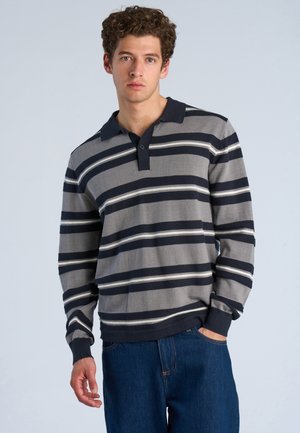 Junger Mann mit lockigem Haar, der ein grau-navy gestreiftes langärmeliges Poloshirt und blaue Jeans vor einem schlichten hellen Hintergrund trägt.